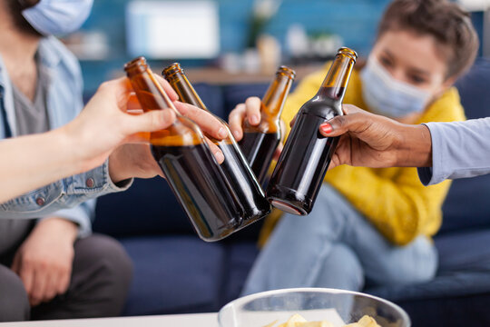 Close up shot of mixed race friends clinking beer bottles hanging out keeping social distancing having fun in apartment linving room during global pandemic. Conceptual image.