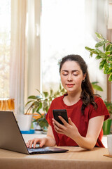 Freelance.A young, pretty woman is working at a laptop and reading a message on her phone. Home decor, the sun outside the window.The concept of quarantine, self-isolation and remote work