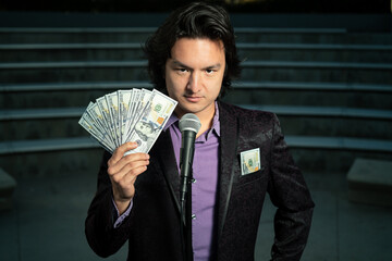 Male Asian Stand Up Comedian with Microphone and Fanned $100 Dollar Bills in Suit in Empty Auditorium Funny Look Face How to Making Money MLM Rich