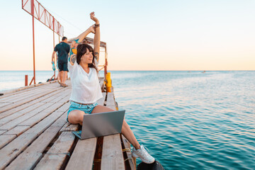 A happy woman with a tattoo on her arm sits on a pier by the sea, and works on a laptop. The concept of remote work and freelancing