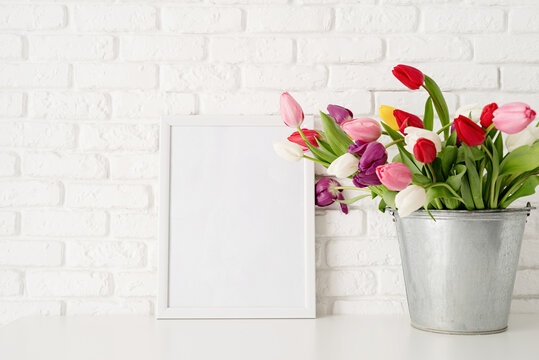 Bucket of fresh tulip flowers and blank frame over white brick wall background