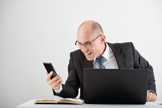 Gloating businessman in glasses sitting at office desk with opened laptop and reading news on failure of business rivals
