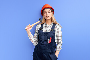 serious constructor woman holding hammer, dressed in overalls and looking at camera, ready to repair broken things, isolated over blue studio background