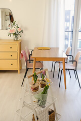 Cozy open dining room and living room interior with a window and a table with chairs. Spring flower decor on a kitchen rolling utility cart. Scandinavian decor concept.