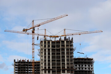 Construction cranes lifting the cargo above unfinished residential buildings on blue sky and white clouds background. Housing construction, apartment block in city