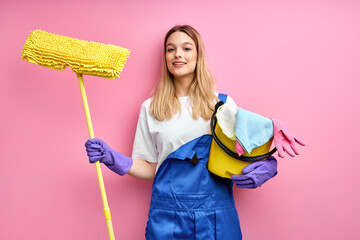 Smiling nice caucasian woman in blue coveralls and rubber gloves look at camera holding cleaning...