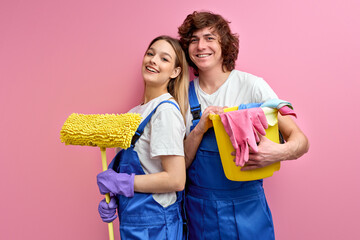 happy couple use rags and other tools for cleaning, posing at camera isolated over pink background. portrait of young man and woman in coveralls excited after household chores, cleaning of home