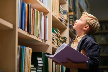 school boy looking up at bookshelves in campus library, child boy holding book in hands