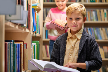 diligent child boy with book between bookshelves in campus library, he is looking at camera....