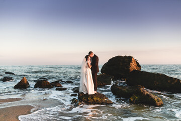 the groom and the bride stand on a stone in the sea at sunset. beautiful wedding couple at sunset...