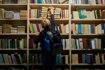 rear view on school boy taking a book from shelves, choosing the book for school, education. intelligent clever children concept