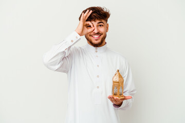 Young Moroccan man wearing a typical arab clothes holding a ramadan lamp isolated on white background excited keeping ok gesture on eye.