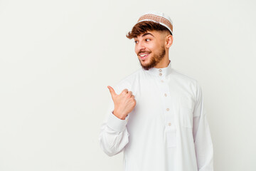 Young Arab man wearing the typical arabic costume isolated on white background points with thumb finger away, laughing and carefree.