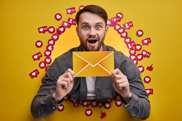 Happy man enjoying positive feedback, want more subscribers and messages, holding letter in hands. Studio portrait of happy white guy among likes on yellow background
