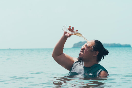 A Funny Asian Guy Pours A Bottle Of Beer On His Head While Standing Chest Deep In The Water At A Beach In The Middle Of The Day.