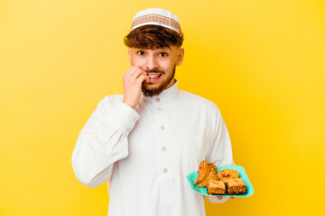 Young Moroccan man wearing the typical arabic costume eating Arabian sweets isolated on yellow background biting fingernails, nervous and very anxious.