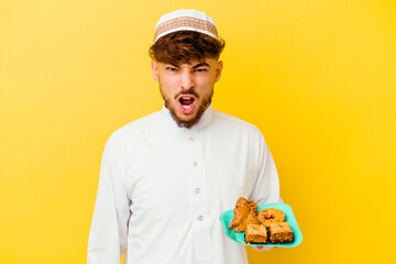 Young Moroccan man wearing the typical arabic costume eating Arabian sweets isolated on yellow background screaming very angry and aggressive.