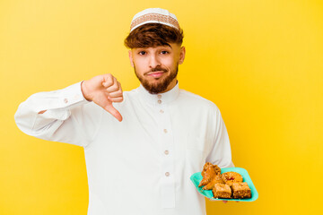 Young Moroccan man wearing the typical arabic costume eating Arabian sweets isolated on yellow background showing a dislike gesture, thumbs down. Disagreement concept.