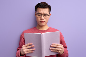 Handsome Chinese Man Reading Book Isolated In Studio , Asian Guy In Eyeglasses Studying, Enjoy Education. Isolated Purple Background