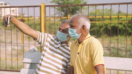 elderly friends with medical face mask taking selfie while sitting at park - concept of happy senior people friendship and meetup after lockdown due to coronavirus covid-19 pandemic
