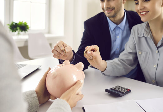 Young Couple Puts Coin In Pink Money Box Sitting At Office Desk During Meeting With Bank Manager, Insurance Broker Or Investment Consultant . People And Finance, Family Budget, Saving Together Concept