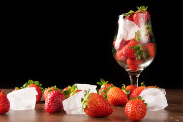 Strawberries, bowl with strawberries and ice and more strawberries and ice cubes on a table, black background, selective focus.