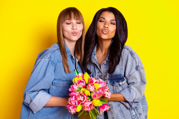 Photo of adorable ladies closed eyes kiss lips arms hold fresh flowers isolated on yellow color background