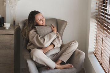 Caucasian woman dressed in comfy grey loungewear sitting in a cozy armchair in her bedroom and holding a decorative pillow. Coziness and comfort concept - 419122236
