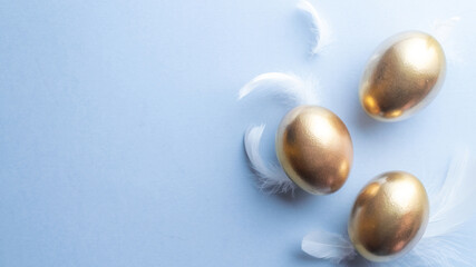 Golden easter colour eggs with white feathers on pastel blue background in Happy Easter decoration. Spring holiday top view concept.