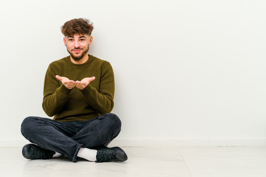 Young Moroccan man sitting on the floor isolated on white background folding lips and holding palms to send air kiss.