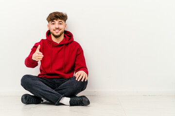 Young Moroccan man sitting on the floor isolated on white background stretching hand at camera in greeting gesture.