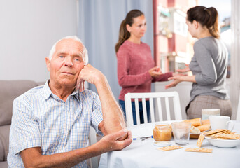 Upset senior man sitting and woman with mother quarreling on background at home