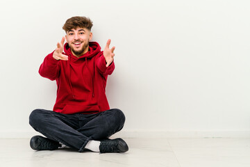 Young Moroccan man sitting on the floor isolated on white background feels confident giving a hug to the camera.