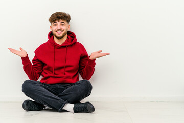 Young Moroccan man sitting on the floor isolated on white background makes scale with arms, feels happy and confident.