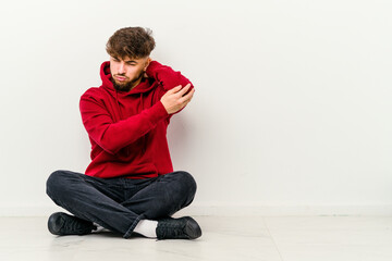 Young Moroccan man sitting on the floor isolated on white background massaging elbow, suffering after a bad movement.