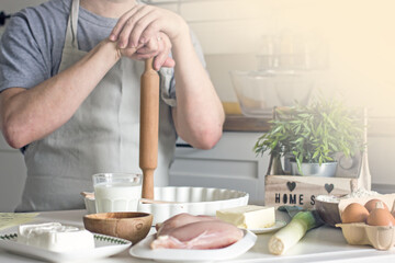 A male cook in an apron holds a rolling pin for rolling out dough in front of the kitchen table with ingredients for the pie. Home cooking an authentic hobby