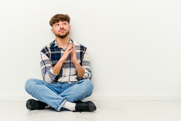 Young Moroccan man sitting on the floor isolated on white background making up plan in mind, setting up an idea.