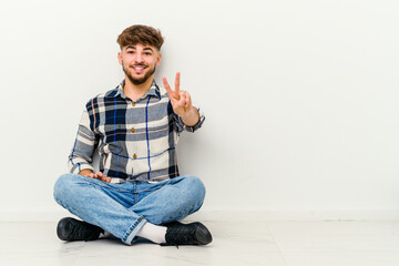 Young Moroccan man sitting on the floor isolated on white background showing victory sign and smiling broadly.