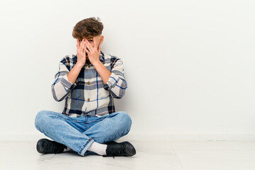 Young Moroccan man sitting on the floor isolated on white background blink through fingers frightened and nervous.