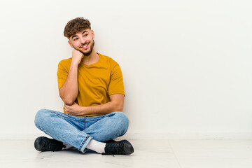 Fototapeta premium Young Moroccan man sitting on the floor isolated on white background smiling happy and confident, touching chin with hand.
