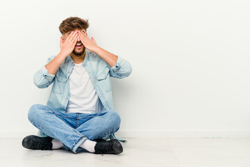 Young Moroccan man sitting on the floor isolated on white background afraid covering eyes with hands.