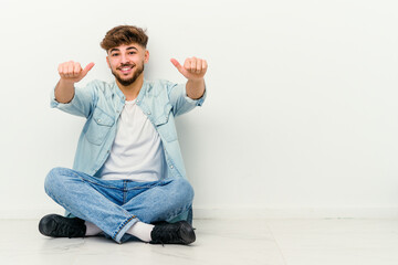Young Moroccan man sitting on the floor isolated on white background raising both thumbs up, smiling and confident.