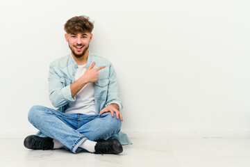 Young Moroccan man sitting on the floor isolated on white background smiling and pointing aside, showing something at blank space.