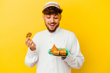 Young Moroccan man wearing the typical arabic costume eating Arabian sweets isolated on yellow background