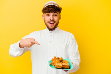 Young Moroccan man wearing the typical arabic costume eating Arabian sweets isolated on yellow background