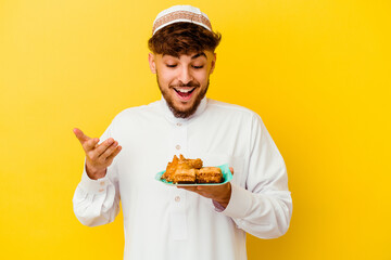 Young Moroccan man wearing the typical arabic costume eating Arabian sweets isolated on yellow background