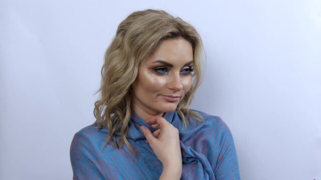 Beautiful young woman with bright evening make-up smoky eyes, freezes and wraps herself in a blue scarf. Girl model thoughtfully looks to the side on a white background.