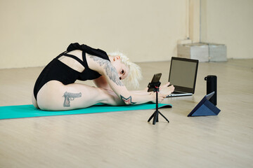 Fit young woman sitting on floor and leaning forward to her legs when filming stretching workout