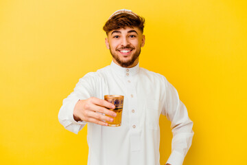 Young Moroccan man wearing the typical arabic costume drinking tea isolated on yellow background
