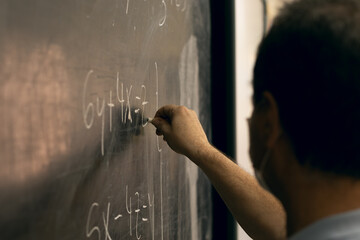 Teacher wearing mask writing equations on a blackboard. Covid situation, pandemic, new normal. Close-up, perspective from the side.